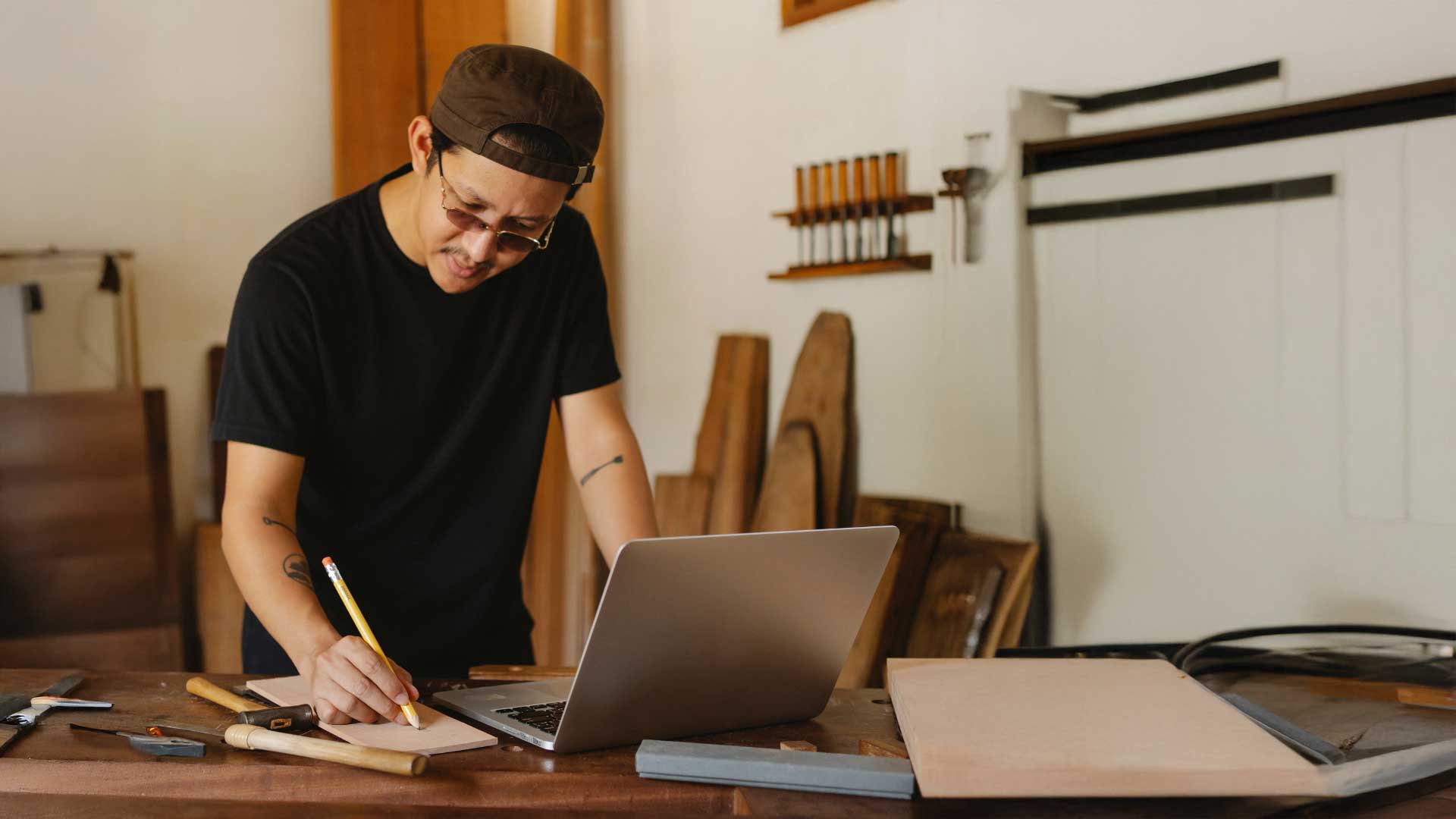 Mund-zu-Mund-Propaganda vs Website: Handwerker mit Kappe und Sonnenbrille steht vor Laptop am Werkbank und notiert etwas mit einem Bleistift.